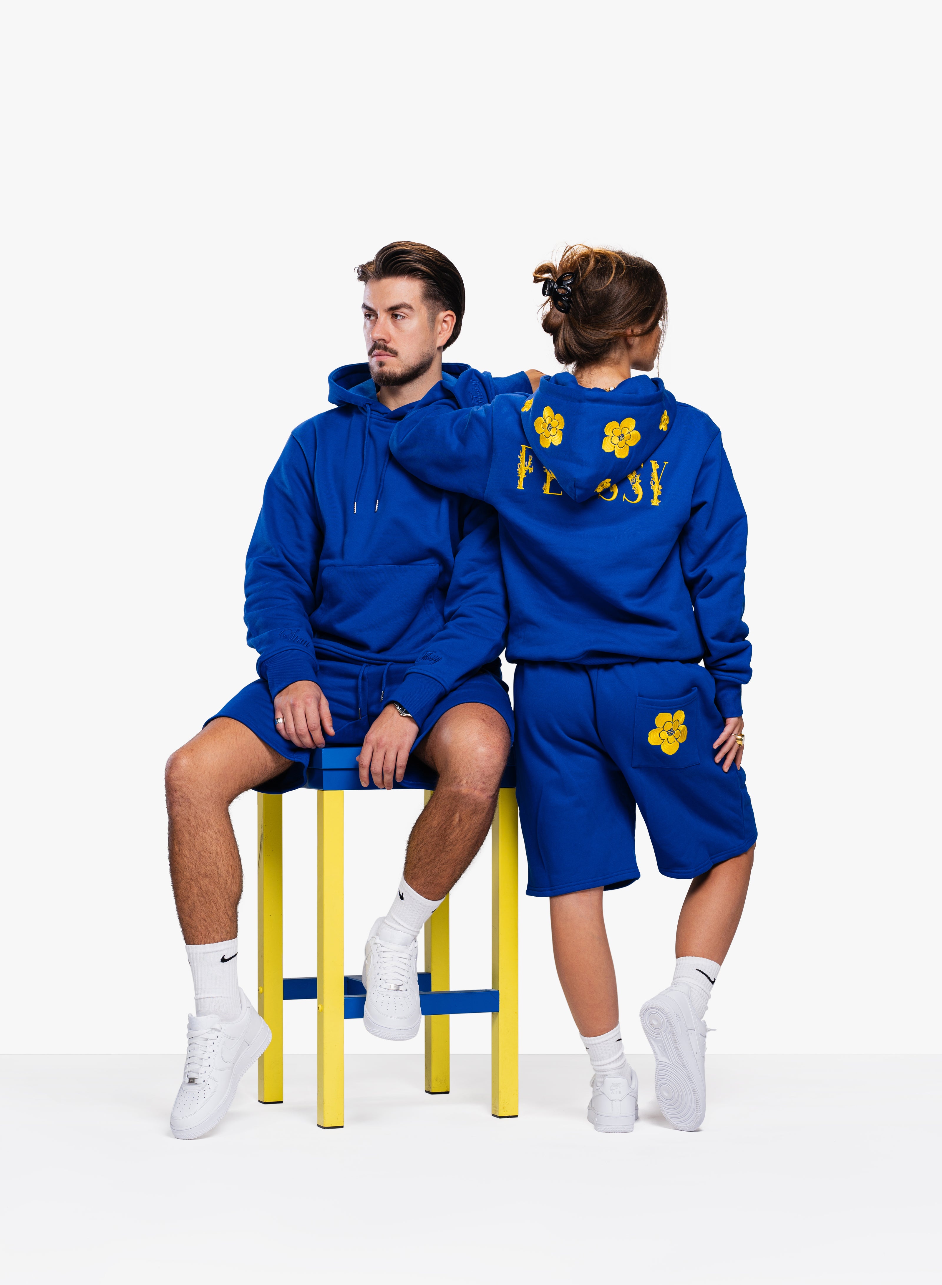 Couple wearing flossy blue hoodies and shorts with yellow floral embroidery, showcasing minimalist and cozy style.